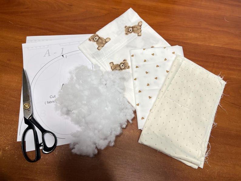 Puede incluir: Un proyecto de manualidades con trozos de tela estampados con osos y corazones, relleno blanco, una hoja de patrones y tijeras. La tela es de color crema con detalles marrones. Las tijeras son negras y plateadas.