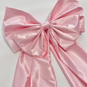 Pode incluir: Um laço grande cor-de-rosa claro feito de um tecido liso e brilhante. O laço apresenta um nó central com duas alças largas e plissadas que se estendem para fora e duas longas caudas de tecido que pendem para baixo a partir do centro.