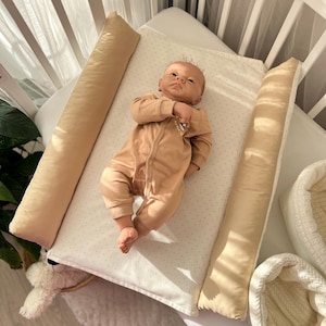 Könnte beinhalten: Eine Babypuppe in einem beigen Langarm-Strampler liegt auf einer rechteckigen Wickelauflage mit weiß-beigem Punktemuster. Die Auflage verfügt über zwei lange, einfarbig beige Seitenpolster zur Stützung und befindet sich in einem weißen Holzbett.