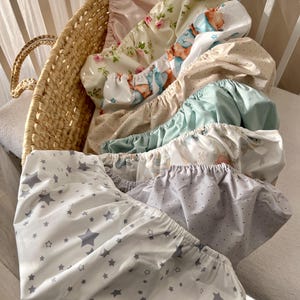 May include: A collection of fitted crib sheets in a woven basket. The sheets feature various patterns, including stars, polka dots, and floral designs. Colors include white, gray, pink, and blue. The sheets are made of soft fabric.