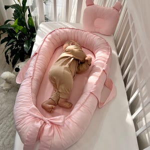 Puede incluir: Un bebé con un mono beige está acostado en un nido rosa para bebés con una almohada rosa a juego en una cuna blanca. El nido tiene un detalle de lazo. La cuna está al lado de una ventana con una cortina blanca.