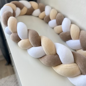 Puede incluir: Un protector de cuna trenzado en una paleta de colores neutros. El parachoques está hecho de tela suave y afelpada y presenta un patrón de secciones blancas, beige y marrones. El diseño es una trenza gruesa y tejida, que proporciona un elemento decorativo y protector para la cuna del bebé.