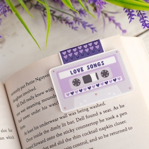 May include: A lavender cassette tape bookmark with the words "LOVE SONGS" printed on it, placed in an open book. The bookmark has a purple heart design. The book is open to Chapter 35.