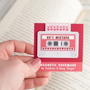 May include: A red magnetic bookmark featuring a retro cassette tape design with the text "80's Mixtape." The bookmark has a white border and is held in a hand. The top of the bookmark has a pattern of pink hearts.