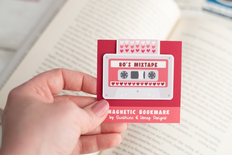 May include: A red magnetic bookmark featuring a retro cassette tape design with the text "80's Mixtape." The bookmark has a white border and is held in a hand. The top of the bookmark has a pattern of pink hearts.