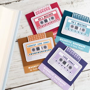 May include: A collection of magnetic bookmarks featuring cassette tape designs. Each bookmark has a different color scheme and text, including "80's Mixtape," "90's Mixtape," "Country Love Songs," and "Love Songs." The bookmarks are on a white wooden surface.