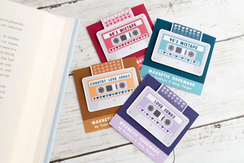 May include: A collection of magnetic bookmarks featuring cassette tape designs. Each bookmark has a different color scheme and text, including "80's Mixtape," "90's Mixtape," "Country Love Songs," and "Love Songs." The bookmarks are on a white wooden surface.