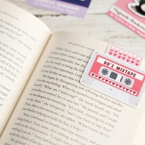May include: A book is open to a page with text, with several magnetic bookmarks. One bookmark is a pink cassette tape design with the text "80's Mixtape". Another bookmark has a pattern of pink hearts.