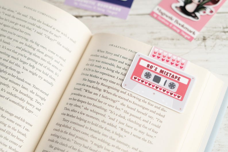 May include: A book is open to a page with text, with several magnetic bookmarks. One bookmark is a pink cassette tape design with the text "80's Mixtape". Another bookmark has a pattern of pink hearts.