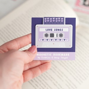 May include: A purple magnetic bookmark featuring a cassette tape design with the words "LOVE SONGS." The bookmark has a purple background with a white border and small purple hearts. The text "MAGNETIC BOOKMARK" is also visible.