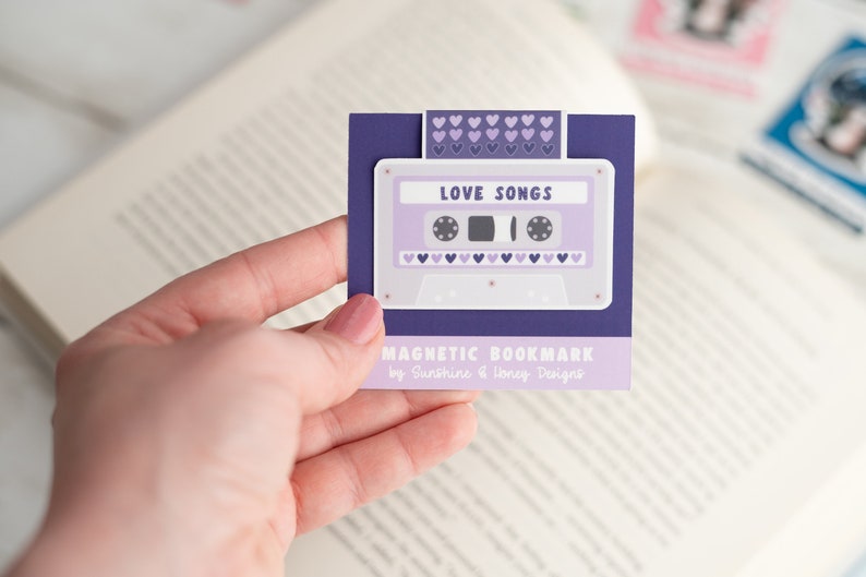 May include: A purple magnetic bookmark featuring a cassette tape design with the words "LOVE SONGS." The bookmark has a purple background with a white border and small purple hearts. The text "MAGNETIC BOOKMARK" is also visible.
