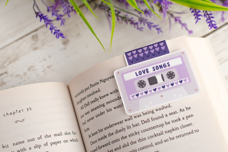 May include: A lavender cassette tape bookmark with the words "LOVE SONGS" printed on it, placed in an open book. The bookmark has a purple heart design. The book is open to Chapter 35.