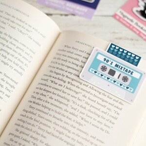 May include: A book is open to a page with text, featuring a light blue and white cassette tape bookmark that reads "90's Mixtape." The bookmark has a row of blue hearts and is placed on the top of the book's pages.