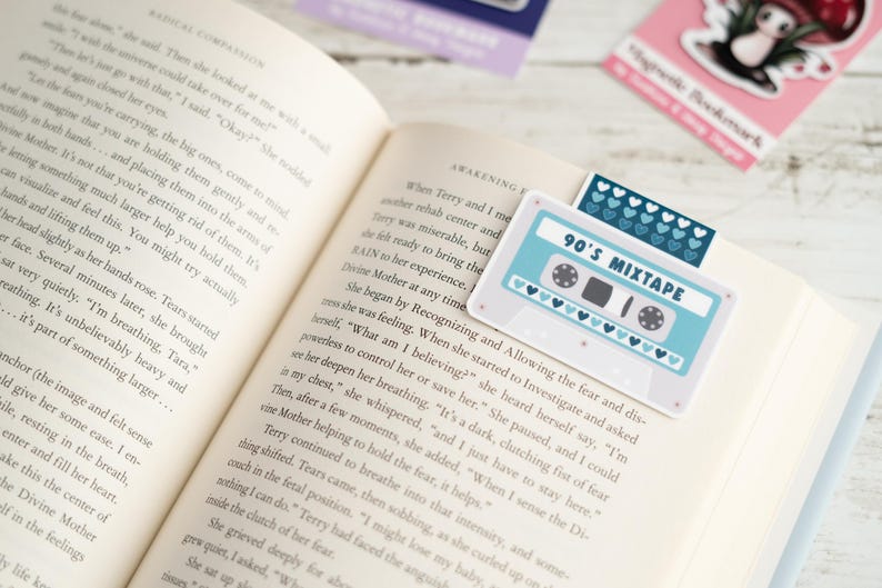 May include: A book is open to a page with text, featuring a light blue and white cassette tape bookmark that reads "90's Mixtape." The bookmark has a row of blue hearts and is placed on the top of the book's pages.