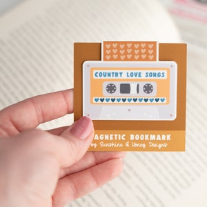 May include: A magnetic bookmark featuring a retro cassette tape design with the words "Country Love Songs." The bookmark is tan with a white cassette tape graphic and blue heart accents. The text "MAGNETIC BOOKMARK" is also visible.