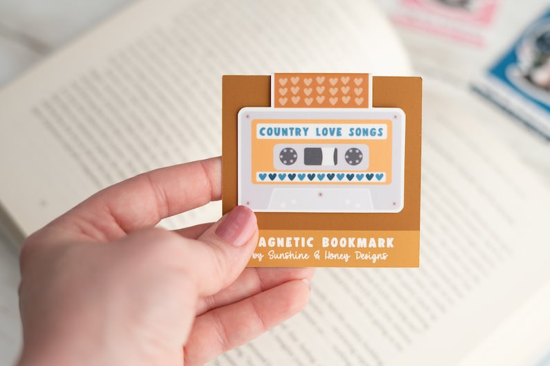 May include: A magnetic bookmark featuring a retro cassette tape design with the words "Country Love Songs." The bookmark is tan with a white cassette tape graphic and blue heart accents. The text "MAGNETIC BOOKMARK" is also visible.