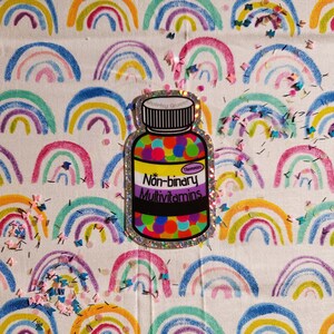May include: A glittery sticker of a pill bottle with the text "Non-binary Multivitamins" and a purple, white, and green flag. The bottle is filled with colorful pills and has a rainbow background.