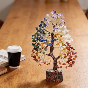 May include: A decorative tree sculpture with a brown trunk and branches, adorned with colorful gemstone leaves. The base is a wooden cylinder. A black coffee cup with a white lid sits to the left.