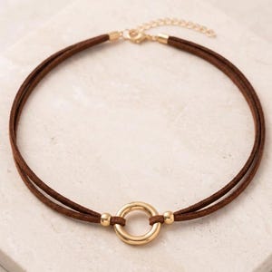 May include: A brown double-strand cord choker necklace featuring a central polished gold-tone metal ring. The necklace includes two small gold-tone metal beads flanking the ring and a gold-tone metal lobster claw clasp with an adjustable chain.