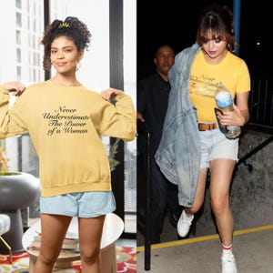 May include: Two women wearing yellow shirts with the text "Never Underestimate The Power of a Woman" printed on them. One woman is wearing a sweatshirt and the other is wearing a t-shirt. Both women are wearing denim shorts.
