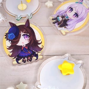 May include: Two circular acrylic pins featuring anime-style characters with purple eyes and hair, one with brown hair and a blue rose hat, the other with lavender hair. The backs show white circular surfaces with yellow star-shaped rubber pin backs.