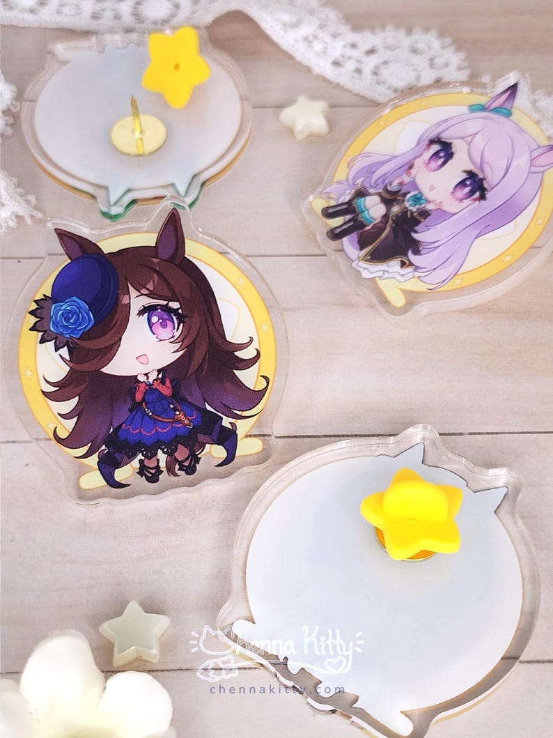 May include: Two circular acrylic pins featuring anime-style characters with purple eyes and hair, one with brown hair and a blue rose hat, the other with lavender hair. The backs show white circular surfaces with yellow star-shaped rubber pin backs.
