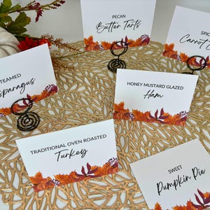 Op de afbeelding: Een set van tien witte kaarten met herfstthema's en zwarte tekst die verschillende Thanksgiving-gerechten opsommen. De kaarten zijn gerangschikt op een gouden placemat met een geweven patroon.