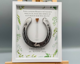 May include: A white frame with a green floral design and a silver horseshoe with a glass vial inside. The text "VON GANZEM HERZEN WÜNSCHEN WIR EUCH, DASS IHR IMMER SO GLÜCKLICH SEID WIE HEUTE!" is printed above the horseshoe. The text "love" is written in cursive below the horseshoe. The text "EMMA & LUKAS 20. AUGUST 2024" is printed below the cursive text. The text "Artina" is printed in the bottom right corner of the frame.