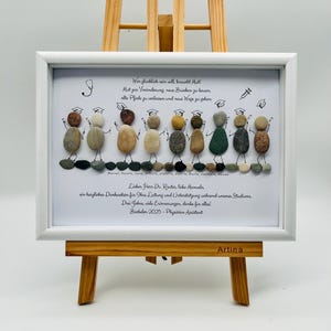 Könnte beinhalten: Ein gerahmtes Kieselkunstwerk mit weißem Rahmen, präsentiert auf einer Holzstaffelei. Die Kunst zeigt Kieselsteinfiguren mit Abschlusskappen und Text auf Deutsch. Der Text enthält motivierende Sätze und eine Dankesbotschaft.