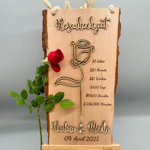 May include: A wooden sign with a rose design and the text "Rosenhochzeit" (German for "Rose Wedding"). The sign also includes the text "10 Jahre", "120 Monate", "521 Wochen", "3.650 Tage", "87.600 Stunden", "5.256.000 Minuten", "Nadine & Michi", "09. April 2025", and "Artina".