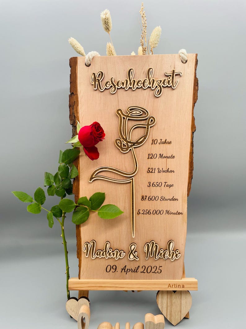 May include: A wooden sign with a rose design and the text "Rosenhochzeit" (German for "Rose Wedding"). The sign also includes the text "10 Jahre", "120 Monate", "521 Wochen", "3.650 Tage", "87.600 Stunden", "5.256.000 Minuten", "Nadine & Michi", "09. April 2025", and "Artina".
