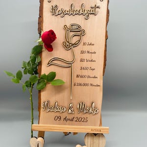 May include: A wooden sign with a rose design and the text "Rosenhochzeit" (German for "Rose Anniversary"). The sign also includes the text "Nadine & Michi" and "09. April 2025". The sign is on a small wooden easel with the word "love" carved in wood.