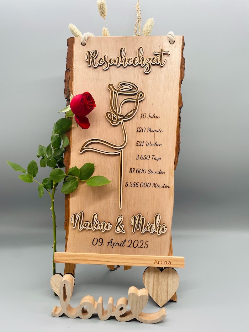 May include: A wooden sign with a rose design and the text "Rosenhochzeit" (German for "Rose Anniversary"). The sign also includes the text "Nadine & Michi" and "09. April 2025". The sign is on a small wooden easel with the word "love" carved in wood.