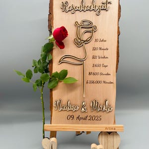 May include: A wooden sign with a carved rose design and the text "Rosenhochzeit" (German for "Rose Wedding"). The sign also includes the text "10 Jahre, 120 Monate, 521 Wochen, 3.650 Tage, 87.600 Stunden, 5.256.000 Minuten" and "Nadine & Michi, 09. April 2025". The sign is on a small wooden easel with the word "love" carved in wood.
