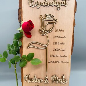 May include: A wooden sign with a carved rose design and the text "Rosenhochzeit" (German for "Rose Wedding"). The sign also includes the text "Nadine & Michi" and "09. April 2025" and the words "10 Jahre", "120 Monate", "521 Wochen", "3.650 Tage", "87.600 Stunden", and "5.256.000 Minuten". The sign is on a small wooden easel.