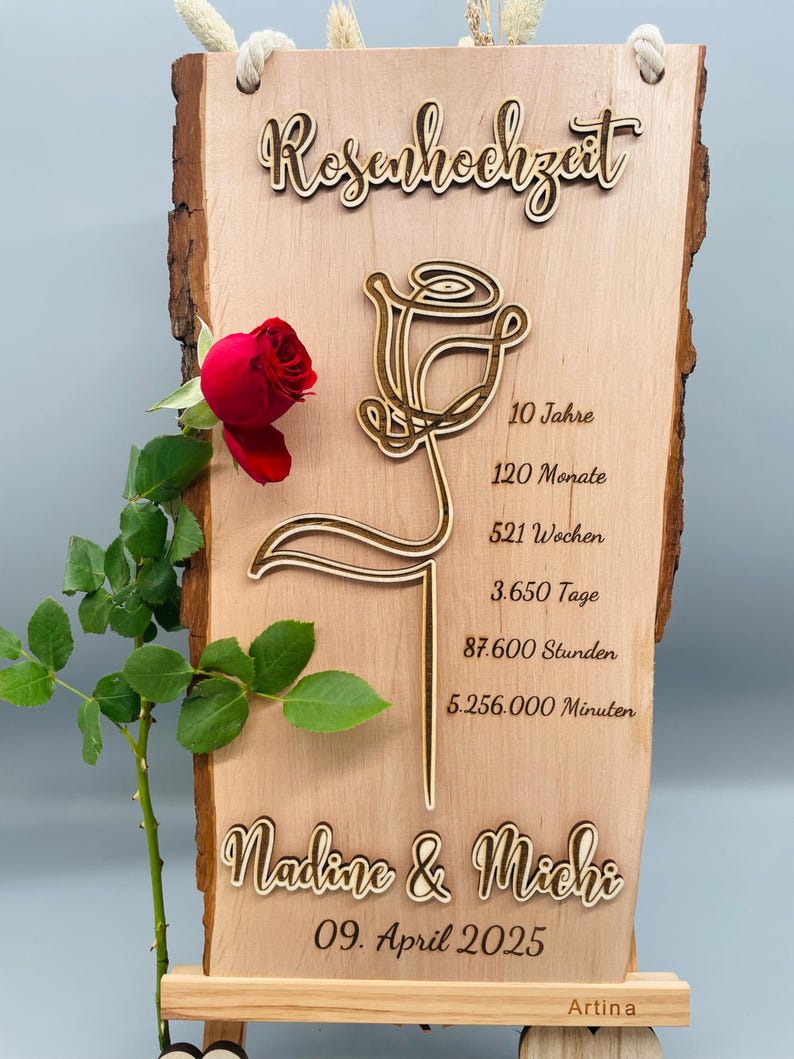 May include: A wooden sign with a carved rose design and the text "Rosenhochzeit" (German for "Rose Wedding"). The sign also includes the text "Nadine & Michi" and "09. April 2025" and the words "10 Jahre", "120 Monate", "521 Wochen", "3.650 Tage", "87.600 Stunden", and "5.256.000 Minuten". The sign is on a small wooden easel.