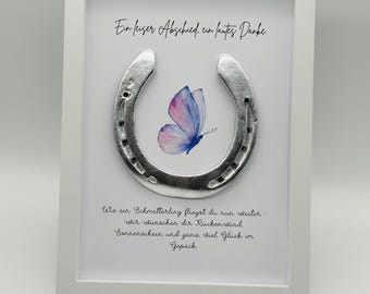 May include: A framed artwork featuring a silver horseshoe and a watercolor butterfly in shades of pink and blue. The artwork includes German text in elegant script. The frame is white.