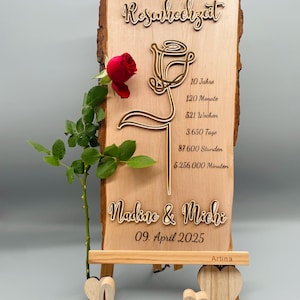 May include: A wooden sign with a carved rose design and the text "Rosenhochzeit" (German for "Rose Wedding"). The sign also includes the text "10 Jahre, 120 Monate, 521 Wochen, 3.650 Tage, 87.600 Stunden, 5.256.000 Minuten" and "Nadine & Michi, 09. April 2025". The sign is on a small wooden easel with the word "love" carved in wood.
