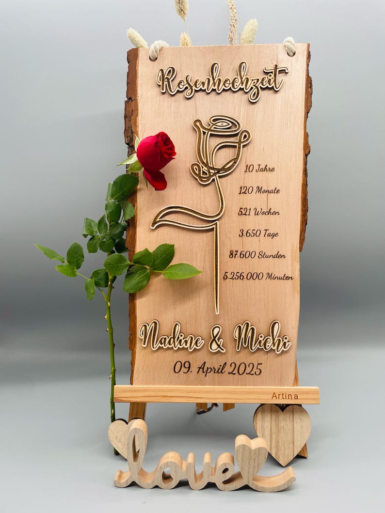 May include: A wooden sign with a carved rose design and the text "Rosenhochzeit" (German for "Rose Wedding"). The sign also includes the text "10 Jahre, 120 Monate, 521 Wochen, 3.650 Tage, 87.600 Stunden, 5.256.000 Minuten" and "Nadine & Michi, 09. April 2025". The sign is on a small wooden easel with the word "love" carved in wood.