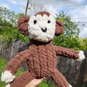 May include: A brown crocheted monkey toy with white accents on the face and feet. The monkey is holding its arms out and has a happy expression.