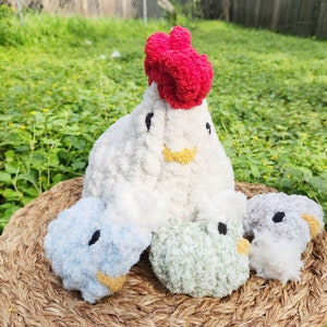 May include: A set of four crocheted stuffed animals shaped like chickens. The main chicken is white with a red comb and yellow beak. The other three chickens are blue, green, and gray.