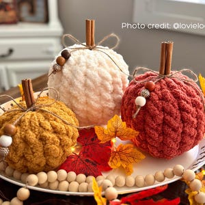 Puede incluir: Tres calabazas hechas a mano con ganchillo en crema, &oacute;xido y amarillo mostaza, cada una con un tallo de canela y un lazo de cuerda. Las calabazas est&aacute;n decoradas con cuentas de madera y se encuentran sobre un plato blanco con hojas de oto&ntilde;o y cuentas de madera.