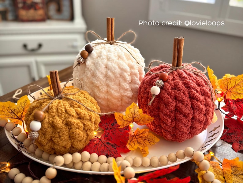 Puede incluir: Tres calabazas hechas a mano con ganchillo en crema, &oacute;xido y amarillo mostaza, cada una con un tallo de canela y un lazo de cuerda. Las calabazas est&aacute;n decoradas con cuentas de madera y se encuentran sobre un plato blanco con hojas de oto&ntilde;o y cuentas de madera.