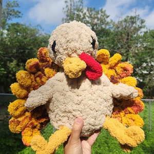 May include: A crocheted turkey plush toy with a beige body, yellow legs, and a red wattle. The turkey has a yellow and orange feathered tail.