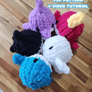 May include: Five handmade crochet baby sea animal toys in various colors: purple, pink, black, white, and blue. The toys have black eyes and are arranged on a wooden surface. Text at the top reads "5IN1 BABY SEA ANIMALS PDF PATTERN + VIDEO TUTORIAL."