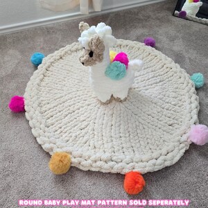 May include: A white crocheted round play mat with colorful pom-poms around the edge. A white crocheted llama toy sits in the center of the mat. The text "ROUND BABY PLAY MAT PATTERN SOLD SEPARATELY @CHUNKYKNITCORNER" is at the bottom of the image.