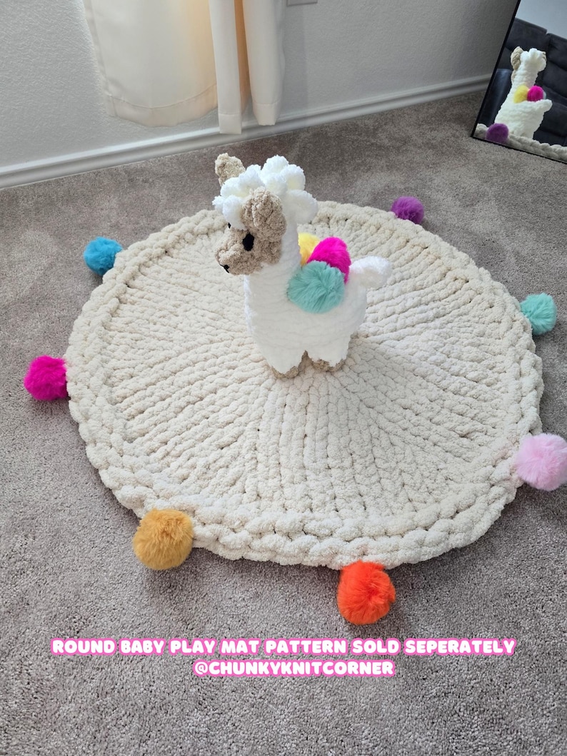 May include: A white crocheted round play mat with colorful pom-poms around the edge. A white crocheted llama toy sits in the center of the mat. The text "ROUND BABY PLAY MAT PATTERN SOLD SEPARATELY @CHUNKYKNITCORNER" is at the bottom of the image.