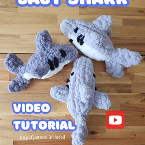 May include: Three grey crocheted baby shark plush toys on a wooden surface. The text "BABY SHARK" is at the top of the image. The text "VIDEO TUTORIAL" is at the bottom of the image. A red play button icon is in the bottom right corner. The text "no pdf pattern included" is below the play button.