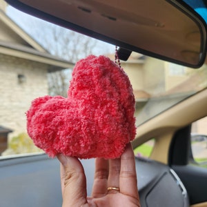 May include: A bright pink heart-shaped plush toy hanging from the rearview mirror of a car.