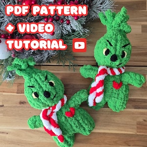 Puede incluir: Dos figuras de ganchillo de color verde que se asemejan al Grinch, con bufandas a rayas rojas y blancas y corazones rojos. Las figuras están sobre una superficie de madera con una corona de Navidad en el fondo. El texto "PDF PATTERN + VIDEO TUTORIAL" es visible sobre las figuras.